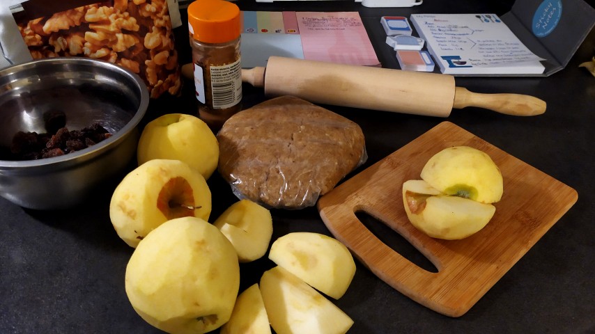 Καθαρισμένα μήλα, ξύλο κοπής, πλάστης, συσκευασία με καρύδια, μπολ με σταφίδες, βαζάκι με κανέλα και σημειωματάριο σε πάγκο κουζίνας