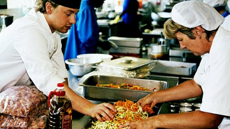 Ο Jamie Oliver και μια γυναίκα ντυμένοι με μαγειρικές στολές, σερβίρουν υγιεινά σχολικά γεύματα