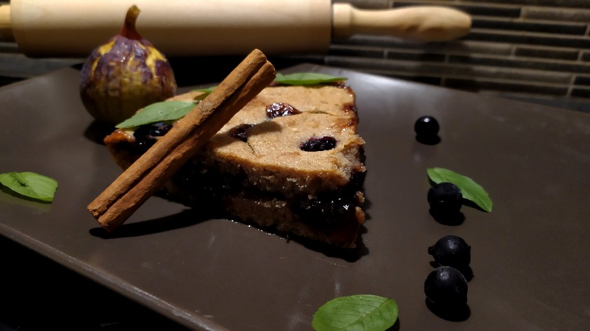 Κομμάτι Linzer torte, ένα στικ κανέλας, ένα σύκο, μερικά φραγκοστάφυλα και φύλλα βασιλικού σε καφέ ορθογώνιο πιάτο, και ένας ξύλινος πλάστης στο βάθος